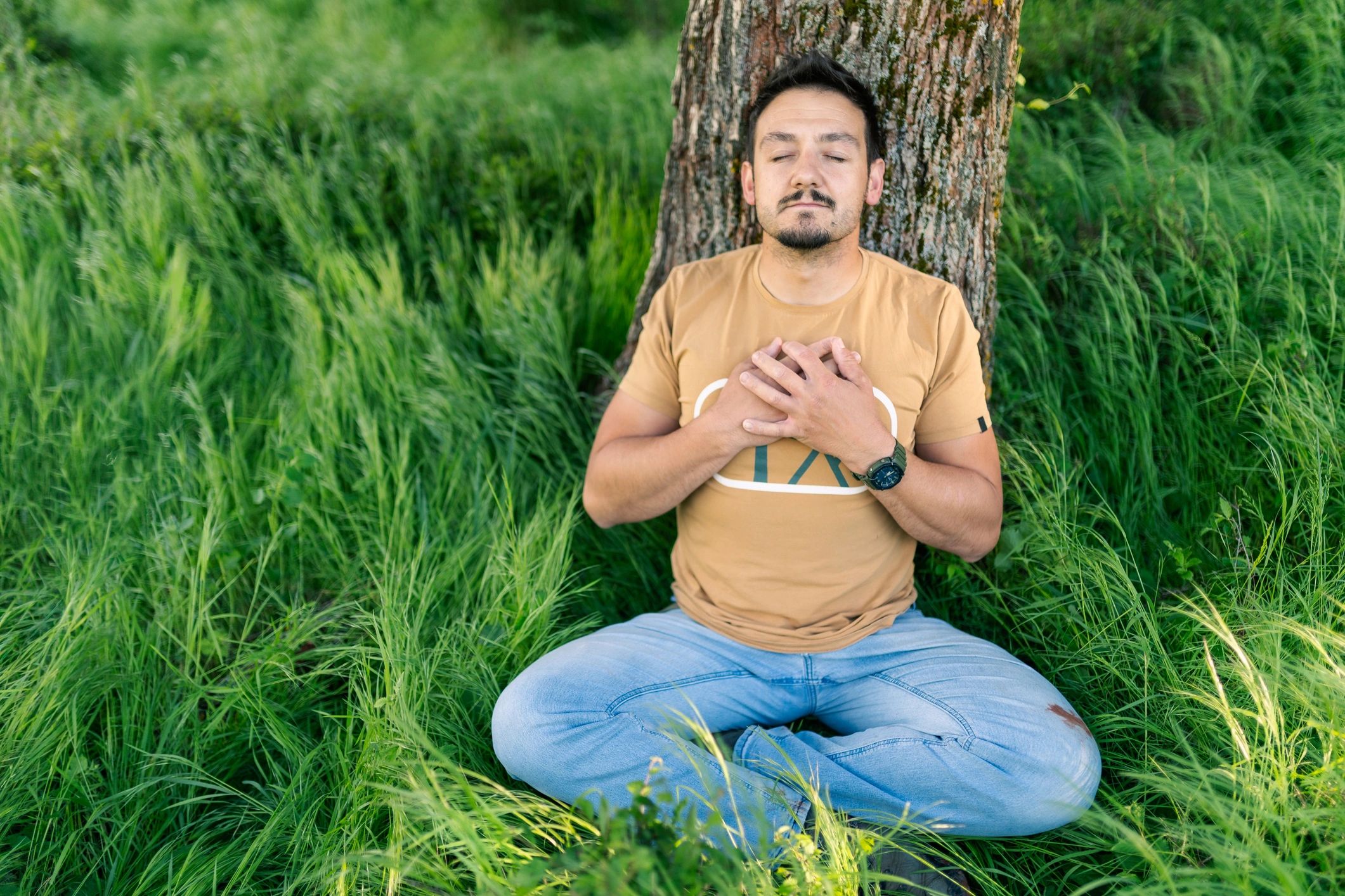 A man with closed eyes is resting in nature, leaning on a tree trunk